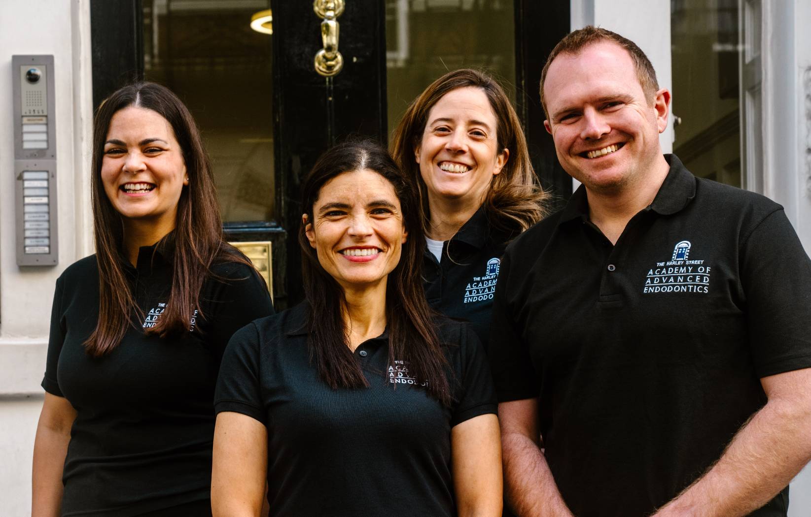 Team image of some of the staff at the Academy of Advanced Endodontics. Team members smiling for the photo in matching black uniform outside of the practice. From right to left- Stella Sarafi, Leigh-Ann Elias, Irene Soriano, Ian Blewitt