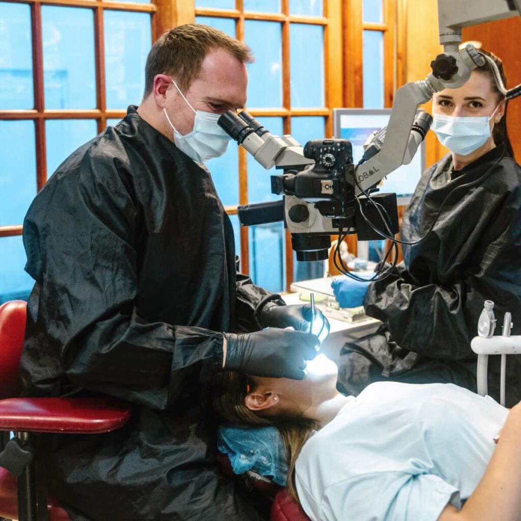 Woman being treated by endodontist in a chair under a dental microscope. Assistant is present in the room and is looking towards the camera.